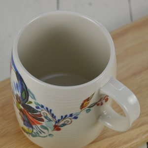 K&ouml;nnte beinhalten: Wei&szlig;e Keramiktasse mit einem bunten Blumenmuster. Die Tasse hat einen breiten, abgerundeten K&ouml;rper und einen Henkel.
