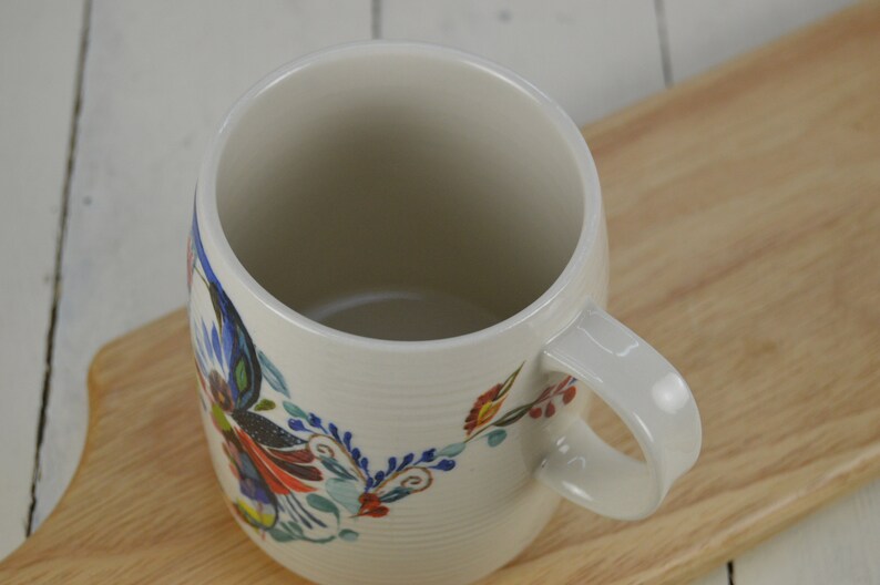 K&ouml;nnte beinhalten: Wei&szlig;e Keramiktasse mit einem bunten Blumenmuster. Die Tasse hat einen breiten, abgerundeten K&ouml;rper und einen Henkel.
