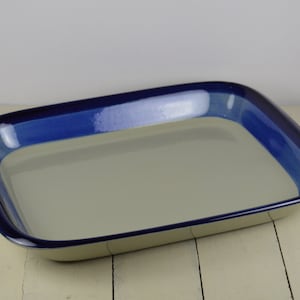 May include: A rectangular ceramic baking dish with a light beige interior and a blue rim. The rim has a darker blue band around the top edge.