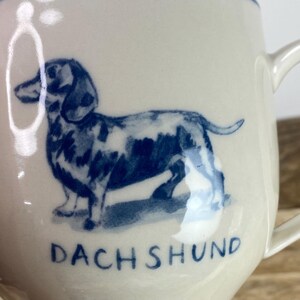 K&ouml;nnte beinhalten: Cremefarbene Keramiktasse mit einer blauen Illustration eines Dackels. Das Wort "DACHSHUND" ist blau unter dem Hund aufgedruckt. Die Tasse hat einen gebogenen Henkel und einen blauen Rand.