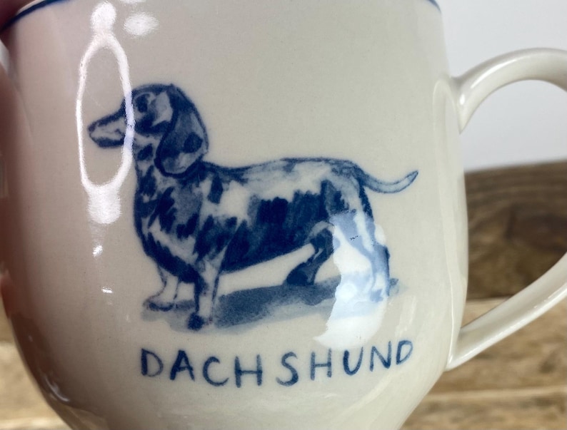 K&ouml;nnte beinhalten: Cremefarbene Keramiktasse mit einer blauen Illustration eines Dackels. Das Wort "DACHSHUND" ist blau unter dem Hund aufgedruckt. Die Tasse hat einen gebogenen Henkel und einen blauen Rand.