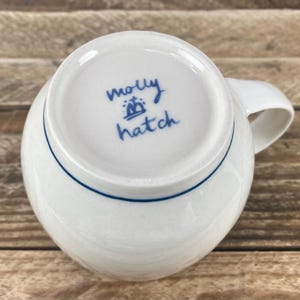 K&ouml;nnte beinhalten: Wei&szlig;e Keramiktasse mit blauem Rand am Boden und gebogenem Henkel. Auf der Unterseite der Tasse steht handschriftlich "molly hatch" in blauer Schrift sowie ein kleines Kronensymbol.