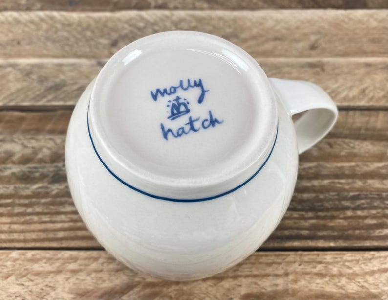 K&ouml;nnte beinhalten: Wei&szlig;e Keramiktasse mit blauem Rand am Boden und gebogenem Henkel. Auf der Unterseite der Tasse steht handschriftlich "molly hatch" in blauer Schrift sowie ein kleines Kronensymbol.