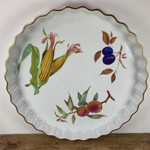 May include: White ceramic serving dish with a scalloped edge and gold trim. The dish is decorated with illustrations of corn, plums, and peaches. The fruits and vegetables are in full color, adding a vibrant touch to the design.