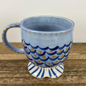 Op de afbeelding: Een lichtblauwe keramische mok met een decoratief golfpatroon in donkerblauw en geel. De handgreep en de rand zijn versierd met gele stippen. De mok staat op een witte voet met blauwe strepen. Een uniek en artistiek stuk.