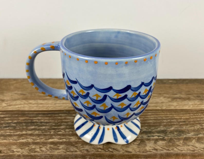 Op de afbeelding: Een lichtblauwe keramische mok met een decoratief golfpatroon in donkerblauw en geel. De handgreep en de rand zijn versierd met gele stippen. De mok staat op een witte voet met blauwe strepen. Een uniek en artistiek stuk.