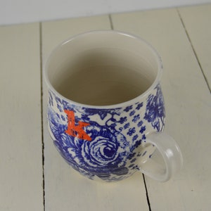 May include: White ceramic mug with a blue floral pattern and an orange letter "K". The mug has a curved shape and a white handle. The design includes roses and other floral elements.