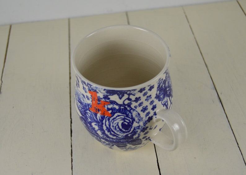May include: White ceramic mug with a blue floral pattern and an orange letter "K". The mug has a curved shape and a white handle. The design includes roses and other floral elements.