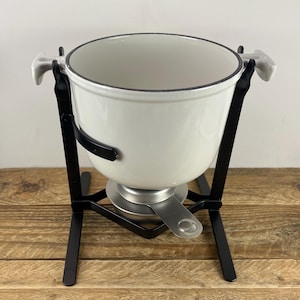 May include: A white fondue pot with a black metal stand. The pot has a dark rim and two white handles. The stand has a circular base with a fuel holder and three legs. The pot is sitting on a wooden surface.