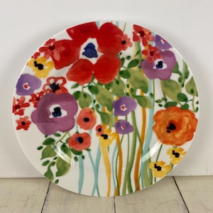 May include: A white ceramic plate with a vibrant floral design. The plate features large red, purple, and orange flowers with green leaves and stems. The flowers are painted in a watercolor style, creating a colorful and artistic look.