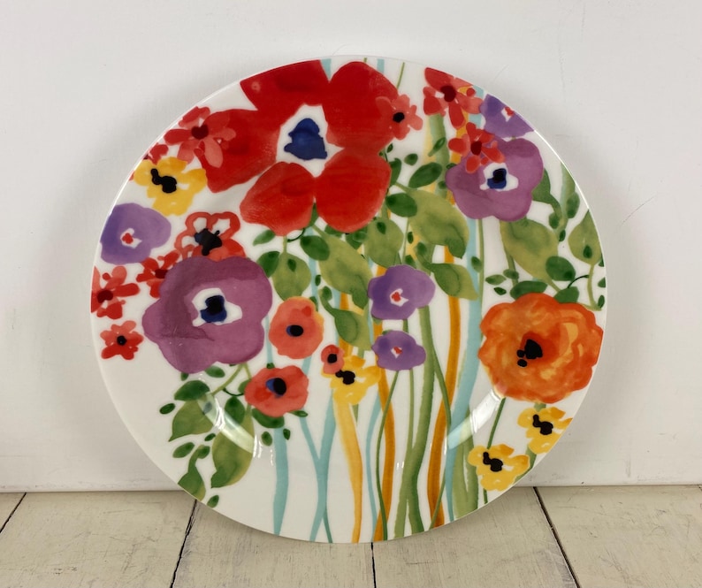 May include: A white ceramic plate with a vibrant floral design. The plate features large red, purple, and orange flowers with green leaves and stems. The flowers are painted in a watercolor style, creating a colorful and artistic look.