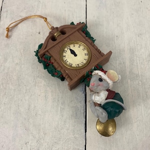 May include: A Christmas ornament featuring a brown cuckoo clock with a gold-colored clock face and a small mouse wearing a Santa hat and green outfit. The mouse is holding onto a gold bell. The ornament has a gold string for hanging.