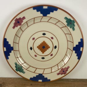 May include: A round ceramic plate with a cream-colored base featuring a central brown diamond shape surrounded by small black and pink geometric accents. The rim displays a repeating pattern of blue, green, and pink stepped shapes and brown lines.