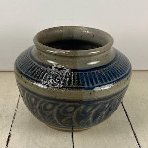 May include: A handcrafted ceramic vase with a wide, rounded body and a slightly flared rim. The vase features a textured design with horizontal blue stripes and a repeating pattern of curved shapes. The color palette includes shades of blue, gray, and brown.