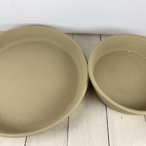 May include: Two beige stoneware baking dishes. One is round and the other is oval. The dishes have a smooth, matte finish and are set on a white wooden surface. These dishes are suitable for baking and serving.