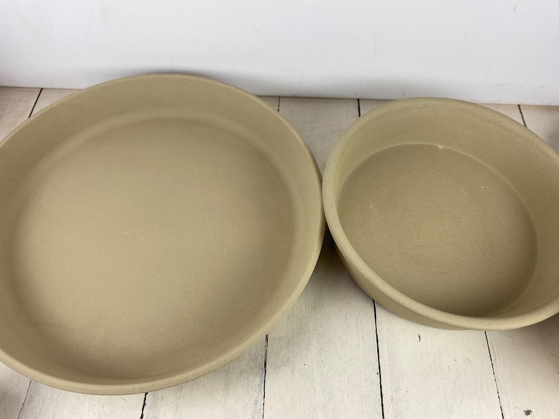 May include: Two beige stoneware baking dishes. One is round and the other is oval. The dishes have a smooth, matte finish and are set on a white wooden surface. These dishes are suitable for baking and serving.