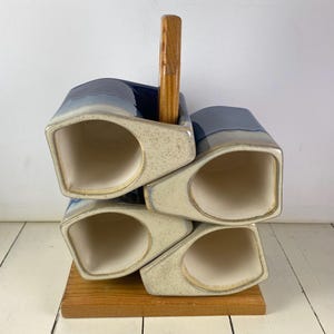 May include: A set of four ceramic mugs arranged in a stack on a wooden stand with a central handle. Each mug features a deep blue glazed upper section that transitions into a speckled cream-colored base with a smooth, off-white interior finish.
