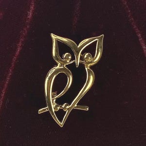 Op de afbeelding: Een goudkleurige uilbroche met een minimalistisch ontwerp. De uil zit op een tak, met een gestileerd lichaam en hoofd. De broche heeft een gepolijste afwerking en staat tegen een donkere achtergrond.