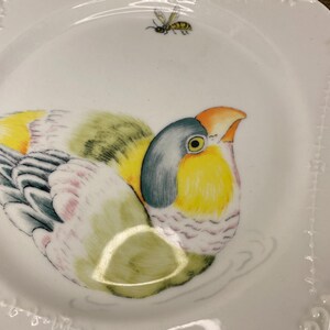 May include: White ceramic plate with a colorful bird illustration. The bird features yellow, green, blue, and orange hues. A small bee is painted above the bird. The plate has a scalloped edge with a delicate embossed design.