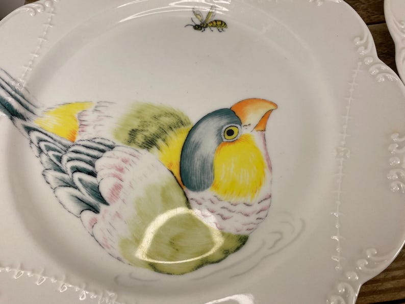 May include: White ceramic plate with a colorful bird illustration. The bird features yellow, green, blue, and orange hues. A small bee is painted above the bird. The plate has a scalloped edge with a delicate embossed design.
