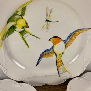 May include: White ceramic plates with a scalloped edge, featuring hand-painted birds and a dragonfly. The birds are depicted in flight with yellow, green, orange, and blue plumage. Decorative tableware for serving.