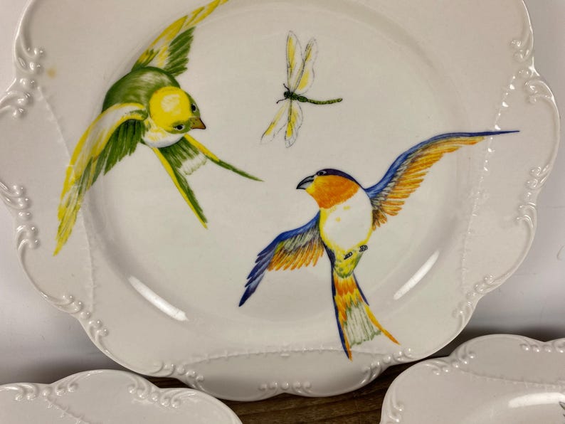 May include: White ceramic plates with a scalloped edge, featuring hand-painted birds and a dragonfly. The birds are depicted in flight with yellow, green, orange, and blue plumage. Decorative tableware for serving.