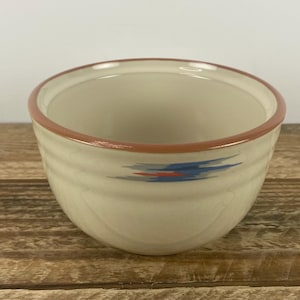 May include: A cream-colored ceramic bowl with a brown rim and a blue and red abstract design. The bowl has a simple, rounded shape and is likely used for serving food or as a decorative item. The bowl is sitting on a wooden surface.