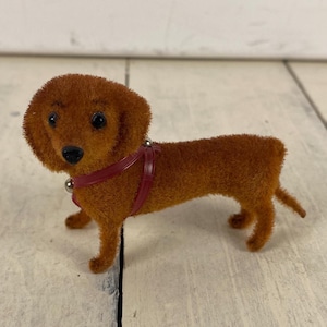 May include: A brown, fuzzy dachshund figurine with a red harness and silver accents. The dog has black eyes and a black nose. The figurine is standing on a light-colored surface.