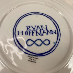K&ouml;nnte beinhalten: Wei&szlig;e Keramikplatte mit blauem kreisf&ouml;rmigen Design mit dem Namen "RYAN HOFFMANN" und einem Unendlichkeitssymbol. Die Platte tr&auml;gt die Aufschriften "Fabriqu&eacute; en Chine" und "hand wash recommended".