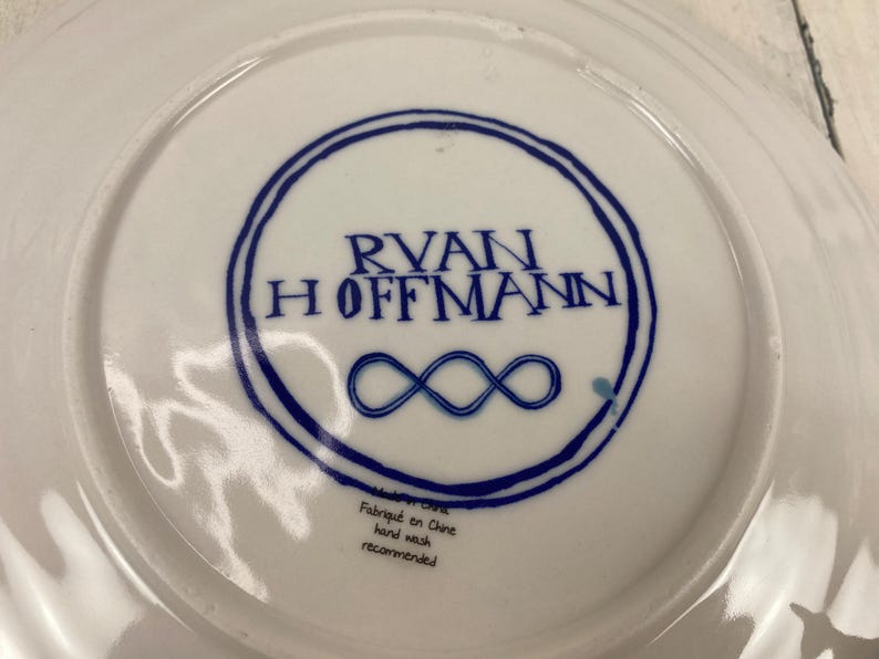 K&ouml;nnte beinhalten: Wei&szlig;e Keramikplatte mit blauem kreisf&ouml;rmigen Design mit dem Namen "RYAN HOFFMANN" und einem Unendlichkeitssymbol. Die Platte tr&auml;gt die Aufschriften "Fabriqu&eacute; en Chine" und "hand wash recommended".
