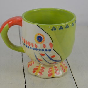May include: A green and white ceramic mug with a colorful floral design. The mug has a handle and a pedestal base.