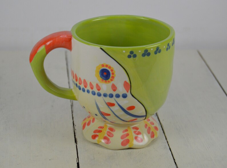 May include: A green and white ceramic mug with a colorful floral design. The mug has a handle and a pedestal base.