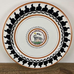 May include: A decorative ceramic plate with a white center, featuring a central emblem of a wolf with two children. The plate has a brown rim, black and white geometric border, and black silhouette accents. A unique piece for display.