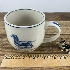 K&ouml;nnte beinhalten: Wei&szlig;er Keramikbecher mit einer blauen Dackel-Illustration und dem Wort "DACHSHUND". Der Becher hat einen blauen Rand und Griffakzente. Eine kleine Hundefigur befindet sich neben dem Becher. Ein Holzlineal ist im Vordergrund.