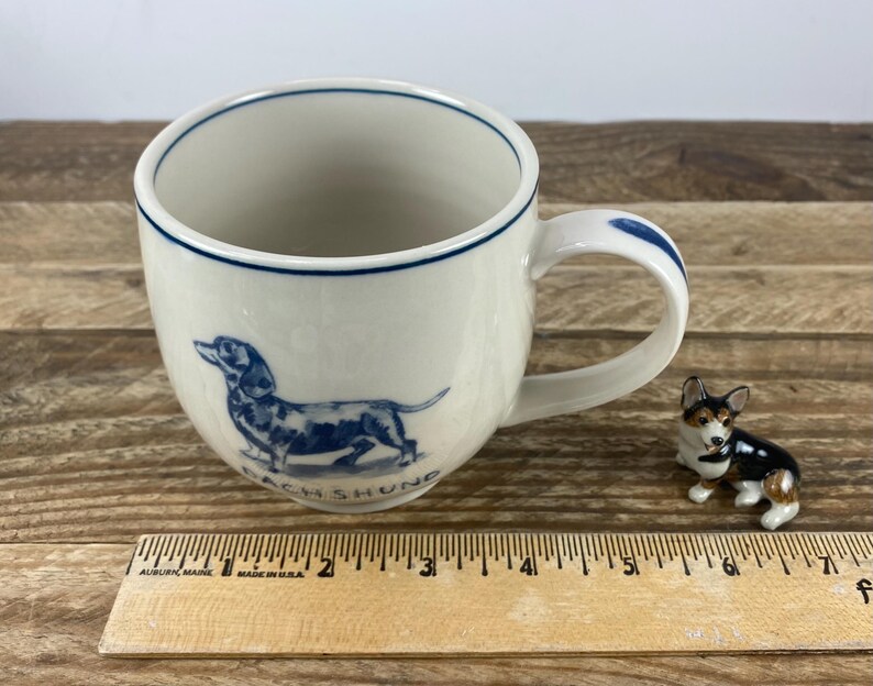 K&ouml;nnte beinhalten: Wei&szlig;er Keramikbecher mit einer blauen Dackel-Illustration und dem Wort "DACHSHUND". Der Becher hat einen blauen Rand und Griffakzente. Eine kleine Hundefigur befindet sich neben dem Becher. Ein Holzlineal ist im Vordergrund.