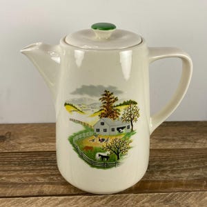 May include: Cream-colored ceramic coffee pot with a lid and handle. The pot features a pastoral scene with a barn, trees, and animals. The lid has a green knob. The pot is on a wooden surface.