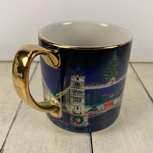 K&ouml;nnte beinhalten: Eine Keramik-Tasse mit goldenem Griff und Rand. Die Tasse zeigt ein n&auml;chtliches Stadtbild-Design mit einem Weihnachtsbaum und einer Br&uuml;cke. Der Hintergrund ist dunkelblau und der Griff hat eine gl&auml;nzende Goldoberfl&auml;che.