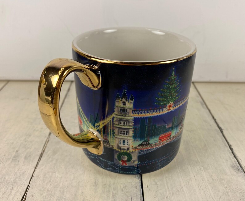 K&ouml;nnte beinhalten: Eine Keramik-Tasse mit goldenem Griff und Rand. Die Tasse zeigt ein n&auml;chtliches Stadtbild-Design mit einem Weihnachtsbaum und einer Br&uuml;cke. Der Hintergrund ist dunkelblau und der Griff hat eine gl&auml;nzende Goldoberfl&auml;che.