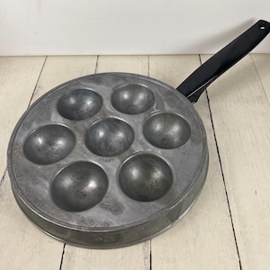 May include: A vintage, round, silver-toned Danish Ebelskiver pan with seven spherical molds. The pan has a black handle and is designed for cooking spherical pancakes. The pan is made of metal and is sitting on a white wooden surface.