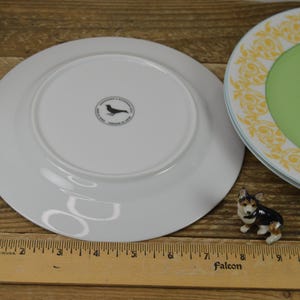 K&ouml;nnte beinhalten: Zwei wei&szlig;e Keramik-Dinnerteller. Ein Teller hat ein schwarzes Vogellogo auf der Unterseite. Der andere Teller hat eine gr&uuml;ne Mitte mit einem gelben und wei&szlig;en Blumenmuster am Rand.
