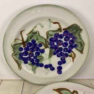May include: Off-white ceramic plate with a green rim, featuring a hand-painted design of purple grapes and green leaves. The grapes are clustered, and the leaves have brown stems. A decorative plate for serving.