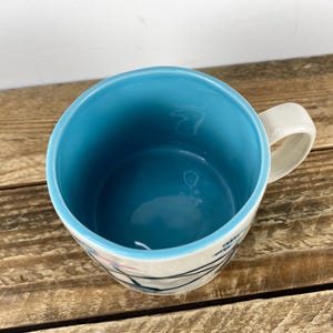 K&ouml;nnte beinhalten: Eine Keramiktasse mit hellblauem Innenraum und wei&szlig;em &Auml;u&szlig;eren. Die Tasse hat einen wei&szlig;en Henkel und ein Design aus schwarzen und rosa Linien. Die Tasse steht auf einer Holzoberfl&auml;che.