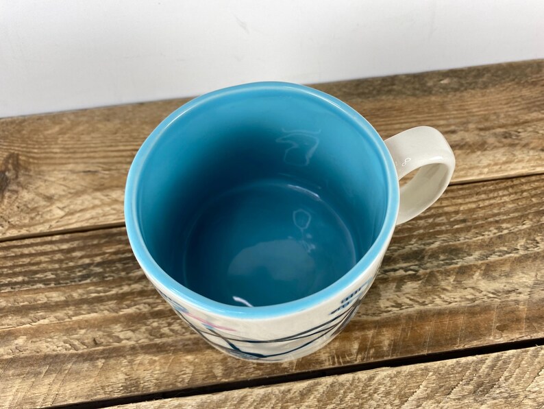 K&ouml;nnte beinhalten: Eine Keramiktasse mit hellblauem Innenraum und wei&szlig;em &Auml;u&szlig;eren. Die Tasse hat einen wei&szlig;en Henkel und ein Design aus schwarzen und rosa Linien. Die Tasse steht auf einer Holzoberfl&auml;che.