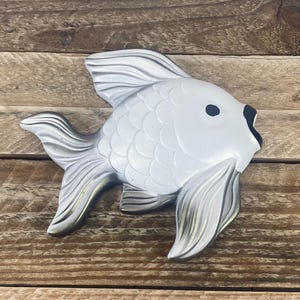 May include: A decorative fish sculpture in shades of white and silver. The fish has detailed fins and scales, with a black eye and mouth. The sculpture is placed on a wooden surface.