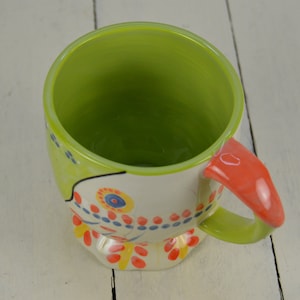 May include: A colorful ceramic mug with a green interior and a whimsical painted design featuring flowers, dots, and swirls. The mug has a red handle.