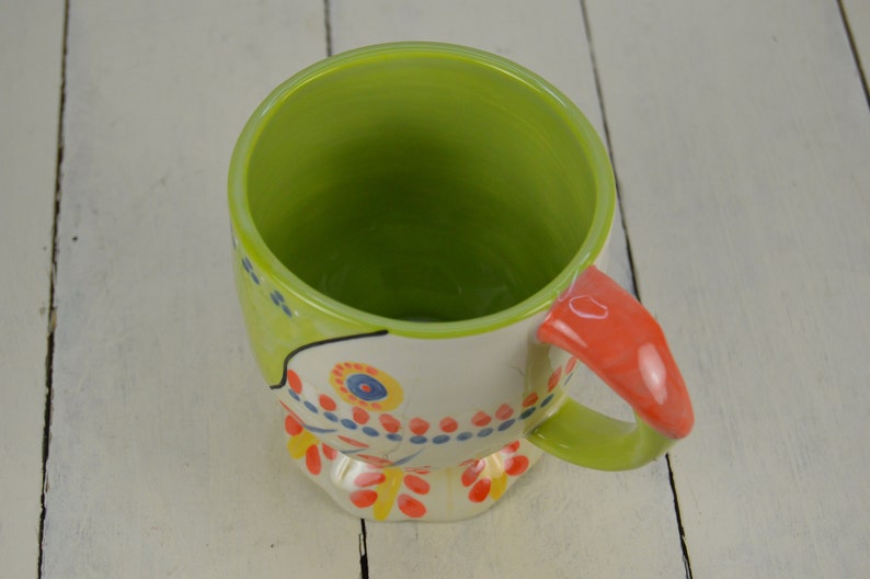 May include: A colorful ceramic mug with a green interior and a whimsical painted design featuring flowers, dots, and swirls. The mug has a red handle.