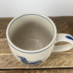 K&ouml;nnte beinhalten: Wei&szlig;er Keramikbecher mit einer blauen Dackel-Illustration. Der Becher hat einen blauen Rand und einen blauen Akzent am Griff. Der Becher steht auf einer Holzoberfl&auml;che.