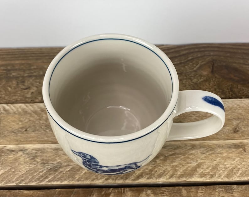 K&ouml;nnte beinhalten: Wei&szlig;er Keramikbecher mit einer blauen Dackel-Illustration. Der Becher hat einen blauen Rand und einen blauen Akzent am Griff. Der Becher steht auf einer Holzoberfl&auml;che.
