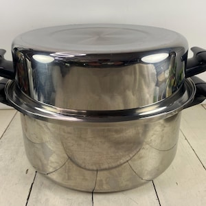 May include: Stainless steel cooking pot set with a lid. The pots are silver with black handles. The set is on a white wooden surface. The pot set is designed for cooking.