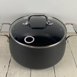 May include: A dark gray cooking pot with a glass lid and stainless steel handles. The pot has two side handles and a single handle on the lid. The lid has a white ring on the inside. The pot is sitting on a white wooden surface.